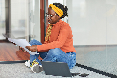 African businesswoman in casual clothing sitting on the floor near the laptop and examining documents at office