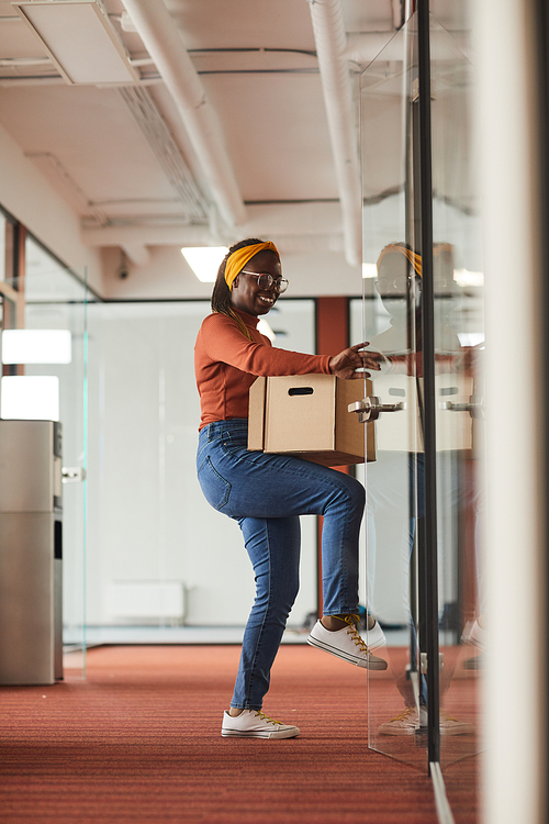 African businesswoman in casual clothing holding box with things and opening the door she is at office