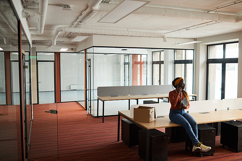 African young businesswoman sitting at the table in empty office and talking on mobile phone