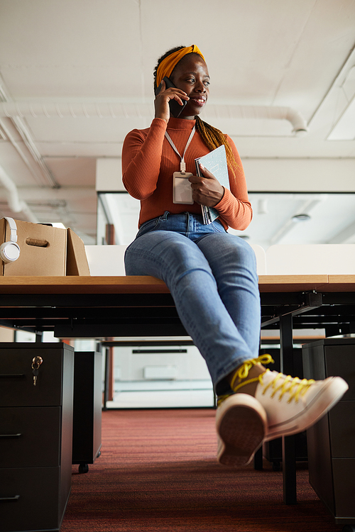 African businesswoman in casual clothing sitting at the table holding documents and talking on her mobile phone at office