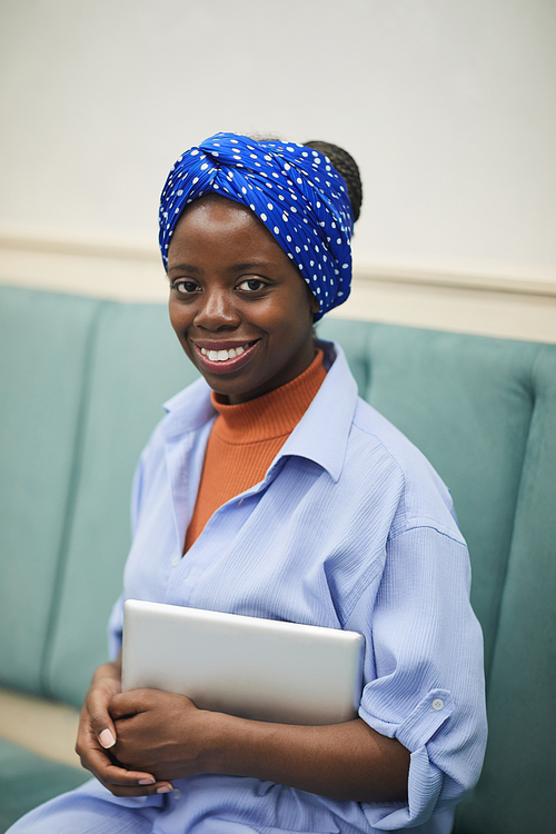 Portrait of African young woman sitting on sofa with digital tablet and smiling at camera