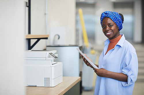 Portrait of African female designer smiling at camera while making copy on copy machine at office