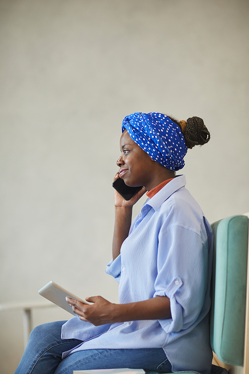 African young businesswoman talking on the mobile phone and using digital tablet while sitting on sofa