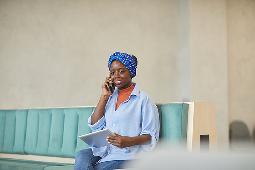 Portrait of African female designer sitting on sofa with tablet pc and has a conversation on the phone with client at office