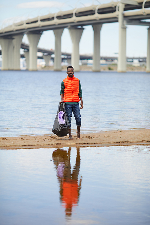 Portrait of African young man standing with garbage on the bank of the river with big bridge in the background