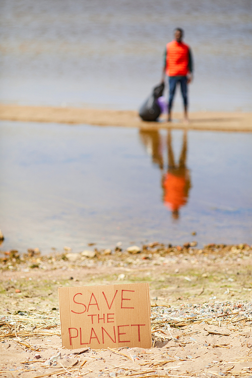 Close-up of cardboard placard with Save the planet on the sand on the bank of the river with man standing in the background