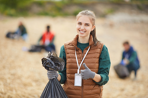 Portrait of young woman holding garbage showing thumb up and smiling at camera while standing outdoors