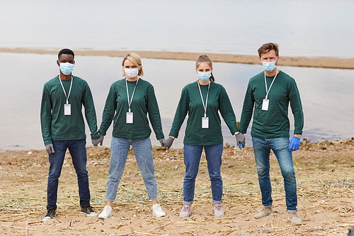 Portrait of group of volunteers in protective masks holding hands while standing on the bank of the river outdoors