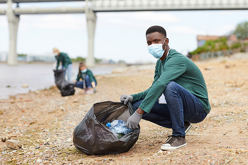 Portrait of African young man in protective mask looking at camera while putting the garbage in the bag on the bank of the river