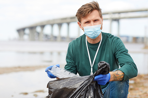Portrait of young volunteer in protective mask putting garbage in bag and looking at camera outdoors