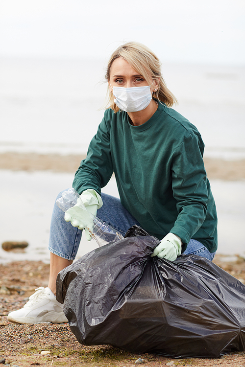 Portrait of young woman in protective mask looking at camera while putting plastic bottles in bag outdoors