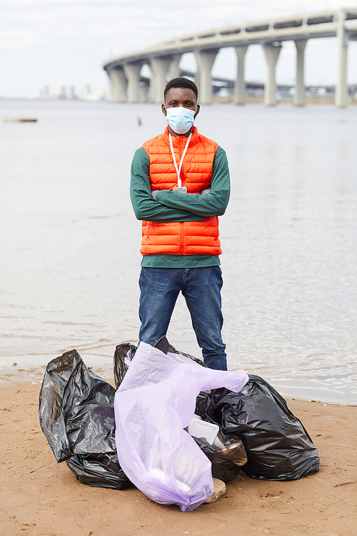 Portrait of African young man in protective mask standing with arms crossed near the rubbish bags near the coastline