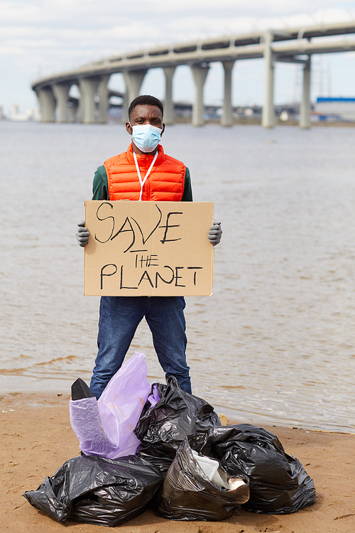 Portrait of African volunteer in protective mask holding placard while standing near the garbage bags near the river
