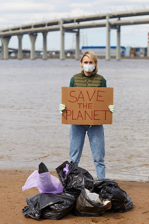 Portrait of young woman in protective mask holding placard Save the planet while standing near the coastline with rubbish bags