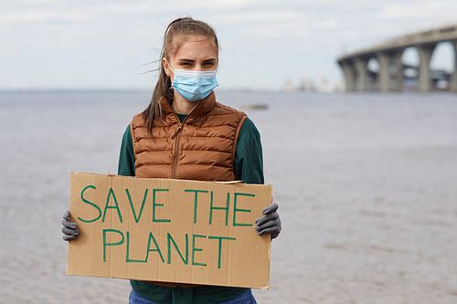 Portrait of young woman in protective mask holding placard Save the planet and looking at camera while standing near the sea