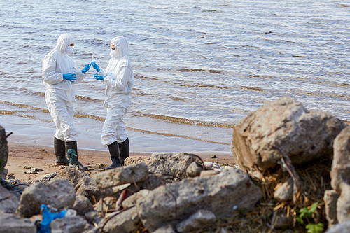 Ecologists in protective suits standing near the coastline and taking samples of water for analysis