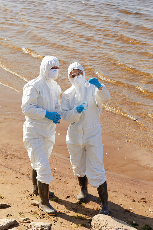 Ecologists in protective suits working in team they standing near the sea holding flask and examining the water in it