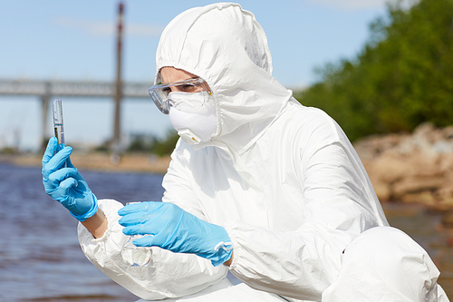 Scientist in protective workwear holding flask with liquid in her hands and examining it outdoors
