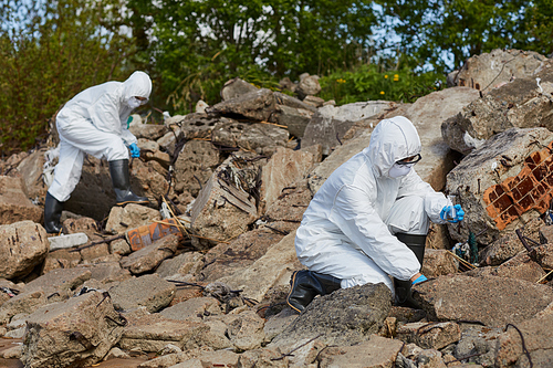 Two colleagues in protective suits holding flasks and taking samples outdoors in rural area