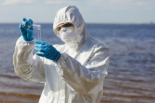 Scientist in protective suit pouring liquid from one flask to another while standing near the sea outdoors
