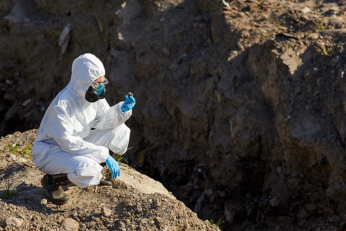 Biologist in protective suit and gloves examining the samples of rocks and stones outdoors