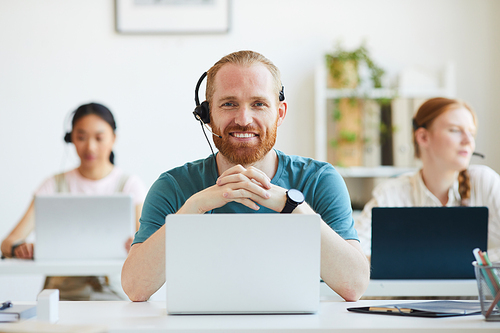 Portrait of bearded operator in headphones smiling at camera while working at the table at office