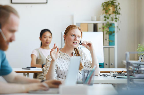 Young woman in headset sitting at her workplace at office and consulting the client on the phone