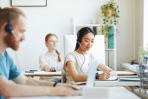 Asian young woman in headphones working at the table together with her colleagues at office