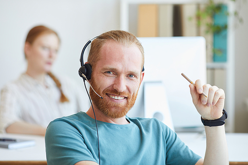 Portrait of young bearded man in headset smiling at camera while working at office