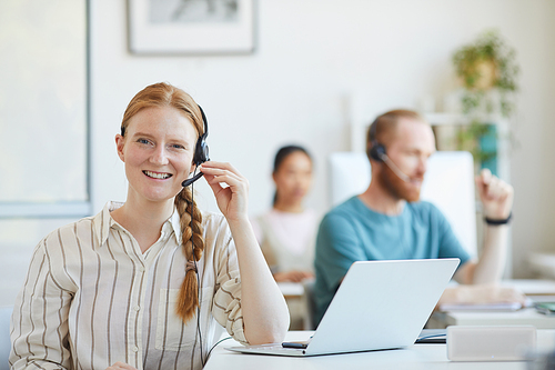 Portrait of young customer service woman in headphones sitting at the table with laptop and smiling at camera at office