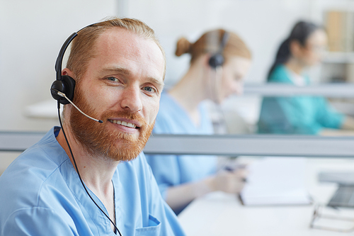 Portrait of bearded young man in headphones smiling at camera while working in call center at office