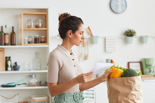 Young woman checking the products from list after shopping while standing in the kitchen