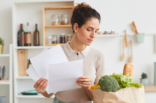Young woman holding documents and checking the products in paper bag while standing in the kitchen