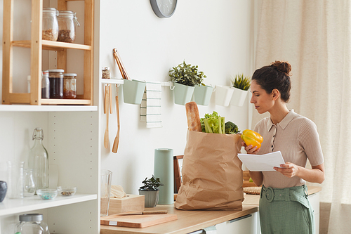 Young woman holding list and looking at vegetables in paper bag while standing in the kitchen