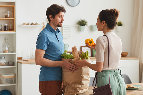 Young couple standing in the kitchen and talking to each other while taking out the vegetables from paper bag