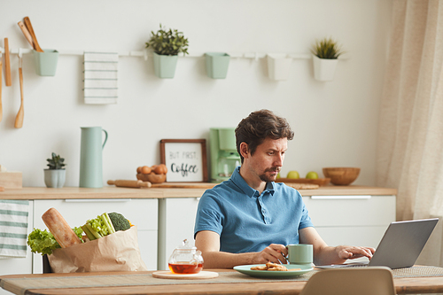 Young man sitting at the table in the kitchen drinking coffee and working online using his laptop