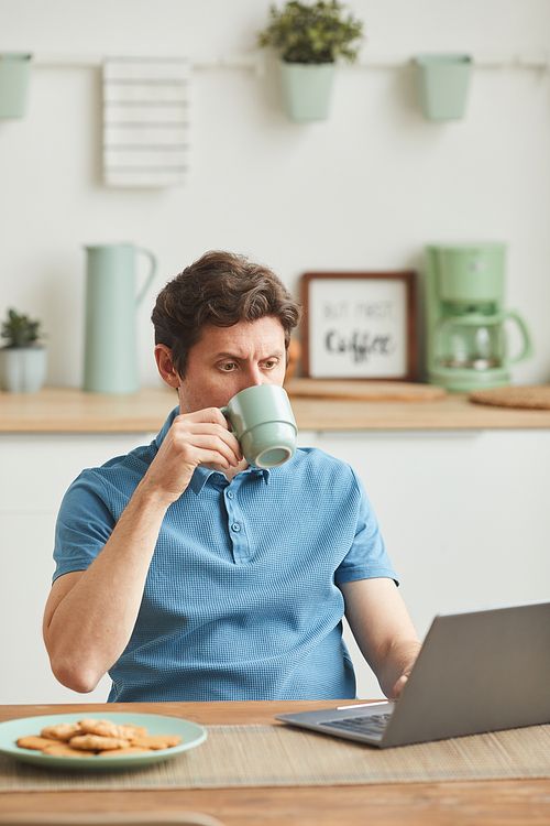 Young man sitting at the table looking at monitor of laptop and drinking coffee in the domestic kitchen