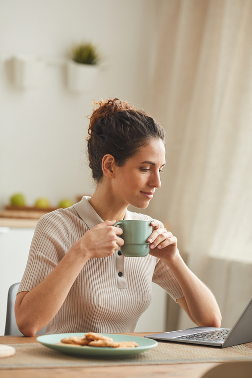 Young woman sitting at the table and drinking coffee while using laptop at home