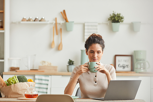 Young woman drinking morning coffee and looking at monitor of laptop while sitting in the kitchen at home