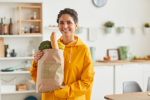 Portrait of young woman holding products in paper bag in her hands and smiling at camera while standing in the domestic kitchen