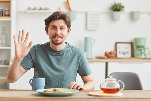 Portrait of young man looking at camera and waving his hand while having breakfast in the kitchen