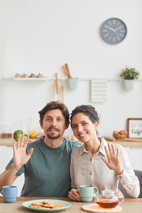 Portrait of young happy couple smiling at camera and waving during their breakfast in the kitchen at home