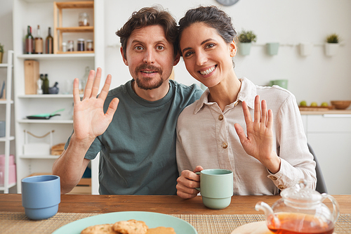 Portrait of young family smiling at camera and waving while sitting at the table and having breakfast in the kitchen