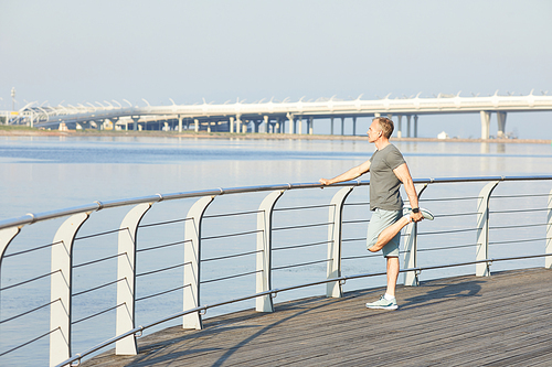 Sporty mature man holding railing and stretching leg on riverside while preparing for running