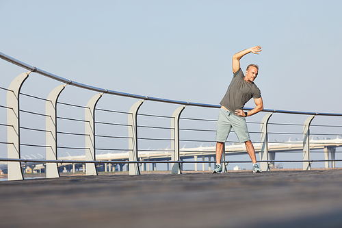 Athletic mature man in sportswear bending side with raised arm while doing warm up on embankment