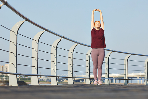 Redhead mature woman in comfortable sportswear raising arms up while doing stretching exercise on embankment