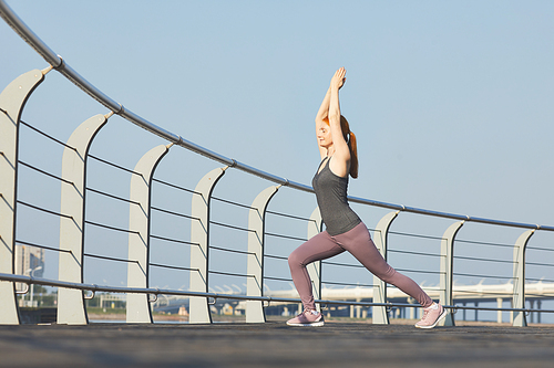 Sporty middle-aged woman with closed eyes performing warrior pose with raised arms on city embankment