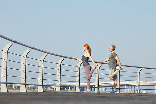 Sporty mature couple standing at railing and stretching legs while training together