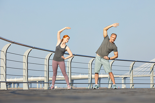 Athletic mature couple standing on city embankment and doing side bend together before training