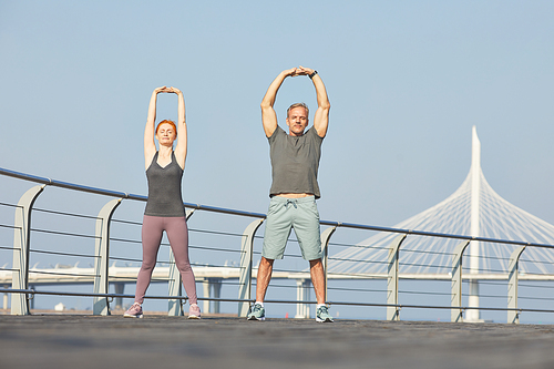 Calm mature couple standing with closed eyes on embankment and strengthening body with fitness exercise